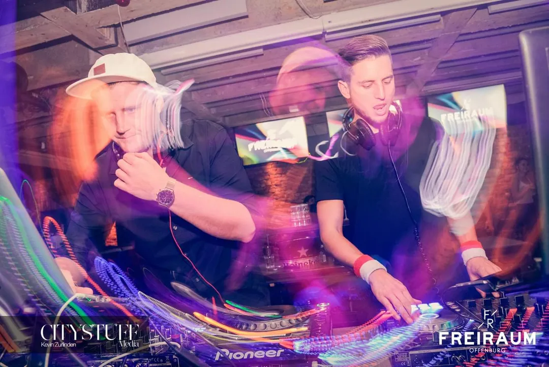 DJ Lukas beim gemeinsamen Auflegen mit einem zweiten DJ an einem Pioneer-Mixpult im Club, umgeben von bunten Lichtstreifen