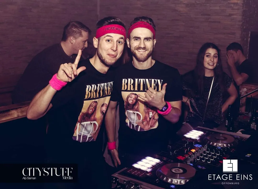DJ Marcel posiert mit einem Kumpel in roten Stirnbändern und Britney-Spears-Shirts vor dem DJ-Pult im Club Etage Eins Offenburg